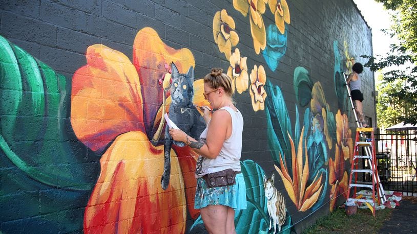 Tiffany Clark (left) and Leslea Hipp (right) of the Mural Machine is creating a mural of cats on the side of Lilyâs Bistro in Daytonâs Oregon District. People who pay a fee will be able to submit pictures of their cats for a realistic paintings on the west side of the restaurant. The project is a fundraiser for local groups dedicated to animal welfare. So far 40 cats are committed to the project and there is room for 85 feline portraits.  LISA POWELL / STAFF